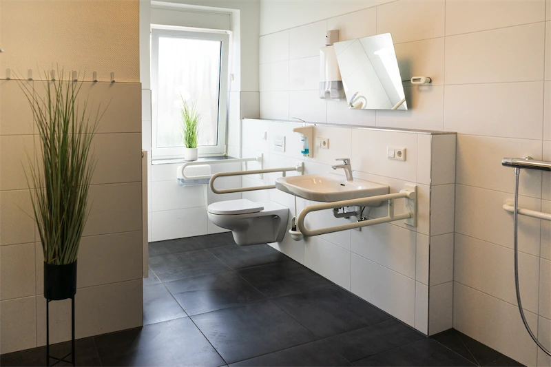 Die Zimmer Barrierefreies Bad im Pflegeheim Bremen Nord: weiße Fliesen, hängendes Waschbecken, WC, Haltegriffe, Fenster, Pflanzen.