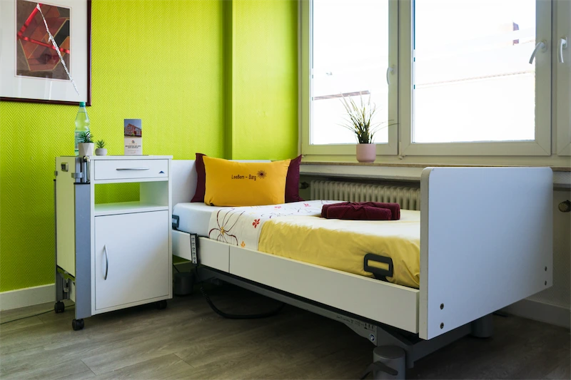 eigene vier Wände Modernes Patientenzimmer im Pflegeheim Bremen Nord mit Einzelbett, gelben Wänden, einem Sideboard, großen Fenstern und Pflanzen.