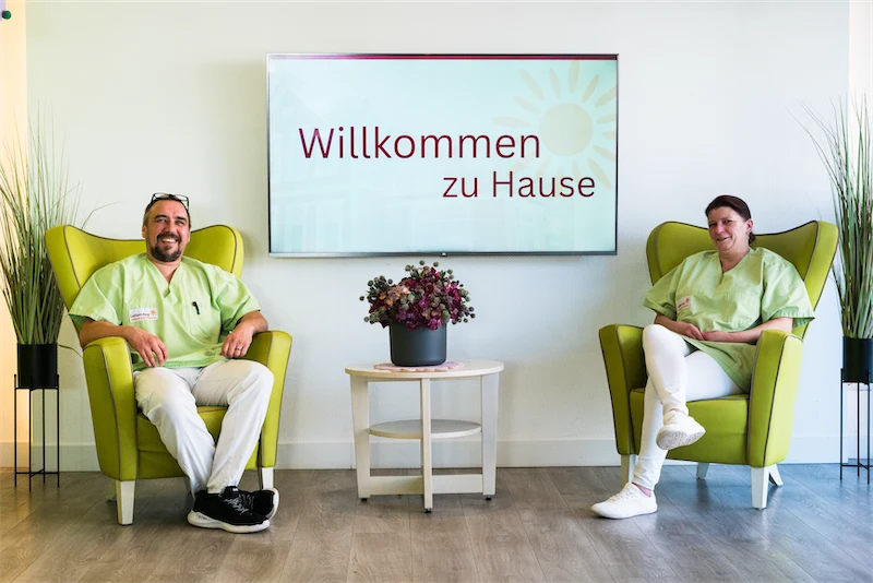 Zwei Personen in grüner Arbeitskleidung sitzen auf limettenfarbenen Sesseln an einem Tisch mit einer Pflanze, auf dem Bildschirm steht: „Willkommen zu Hause“.